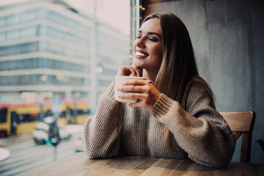 Woman in brown sweater holding cup of coffee looking out window smiling